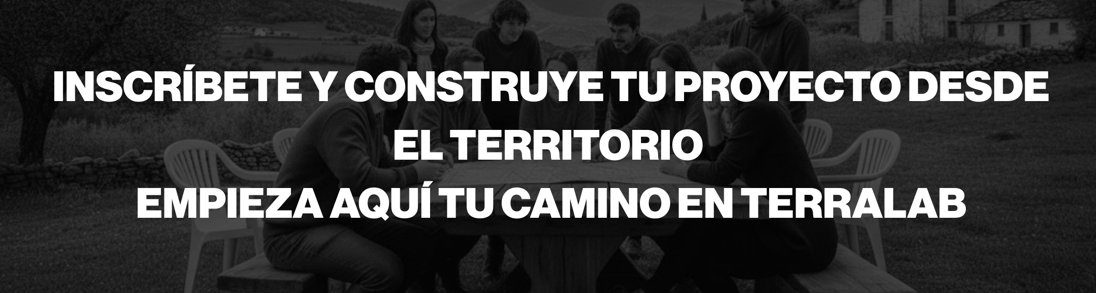 La imagen es una fotografía en blanco y negro de un grupo de personas reunidas alrededor de una mesa rectangular al aire libre, en un entorno rural. Están sentadas en sillas sencillas, conversando y trabajando juntas, lo que transmite colaboración y toma de decisiones compartida. Al fondo se distinguen colinas, campos y construcciones rurales, reforzando la conexión con el territorio. Sobre la imagen aparece un texto grande y contundente en letras blancas: “INSCRÍBETE Y CONSTRUYE TU PROYECTO DESDE EL TERRITORIO. EMPIEZA AQUÍ TU CAMINO EN TERRALAB”. El conjunto evoca comunidad, acción colectiva y un proceso de emprendimiento ligado al entorno rural, con un tono serio, comprometido y participativo.